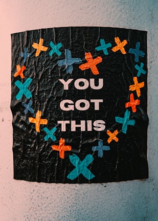 A textured black poster with bright colored crosses forming a heart shape around the words 'You Got This'. The crosses are in shades of blue, orange, and teal, giving a vibrant contrast to the dark background.