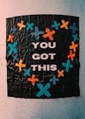A textured black poster with bright colored crosses forming a heart shape around the words 'You Got This'. The crosses are in shades of blue, orange, and teal, giving a vibrant contrast to the dark background.