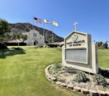A church with a light beige facade featuring a circular design above the main entrance stands prominently. Three flags, including the American flag, are flying on tall poles near the church. The foreground showcases a manicured lawn with a flowering garden encircling a sign that reads 'Saint Francis of Assisi'. A backdrop of mountains adds depth to the scene.