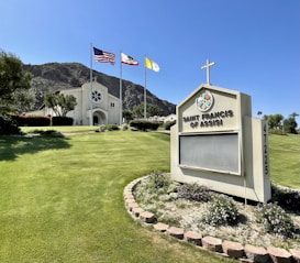 A church with a light beige facade featuring a circular design above the main entrance stands prominently. Three flags, including the American flag, are flying on tall poles near the church. The foreground showcases a manicured lawn with a flowering garden encircling a sign that reads 'Saint Francis of Assisi'. A backdrop of mountains adds depth to the scene.