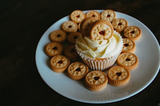 A cupcake with white cream frosting is topped and surrounded by several small round cookies. The cookies have a heart-shaped cutout in the center revealing some red filling. The arrangement is placed on a simple white plate against a dark background.