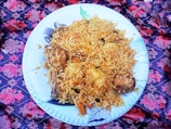 A large white plate filled with a generous serving of biryani rice, garnished with chunks of meat and potatoes. The dish is placed on a vibrant, patterned cloth featuring red and purple floral designs.