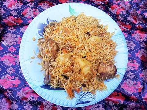 A large white plate filled with a generous serving of biryani rice, garnished with chunks of meat and potatoes. The dish is placed on a vibrant, patterned cloth featuring red and purple floral designs.