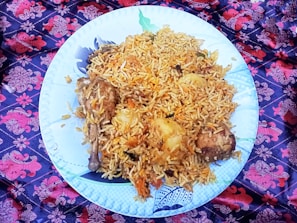 A large white plate filled with a generous serving of biryani rice, garnished with chunks of meat and potatoes. The dish is placed on a vibrant, patterned cloth featuring red and purple floral designs.