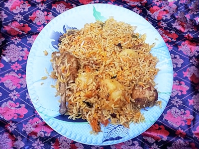 A large white plate filled with a generous serving of biryani rice, garnished with chunks of meat and potatoes. The dish is placed on a vibrant, patterned cloth featuring red and purple floral designs.