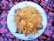 A large white plate filled with a generous serving of biryani rice, garnished with chunks of meat and potatoes. The dish is placed on a vibrant, patterned cloth featuring red and purple floral designs.