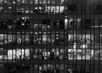 Office building facade at night with illuminated windows, revealing multiple floors and offices inside. Silhouettes of people are visible working late into the night.