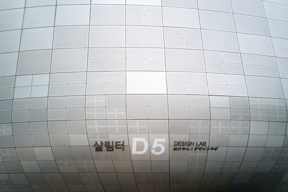 A section of an architectural structure features a pattern of metallic, perforated panels in various sizes. The surface creates a subtle, repetitive grid design, displaying clean lines and geometric precision. Notably, there is signage indicating 'D5 Design Lab' in English and another language.