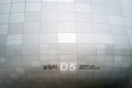 A section of an architectural structure features a pattern of metallic, perforated panels in various sizes. The surface creates a subtle, repetitive grid design, displaying clean lines and geometric precision. Notably, there is signage indicating 'D5 Design Lab' in English and another language.