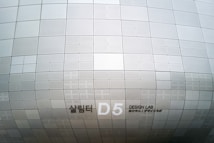 A section of an architectural structure features a pattern of metallic, perforated panels in various sizes. The surface creates a subtle, repetitive grid design, displaying clean lines and geometric precision. Notably, there is signage indicating 'D5 Design Lab' in English and another language.