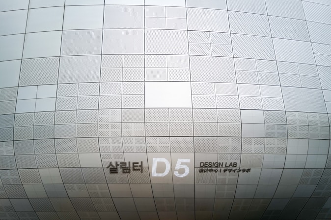 A section of an architectural structure features a pattern of metallic, perforated panels in various sizes. The surface creates a subtle, repetitive grid design, displaying clean lines and geometric precision. Notably, there is signage indicating 'D5 Design Lab' in English and another language.