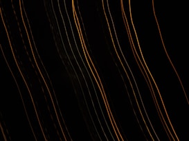 Several curved, parallel lines of yellow and orange light are set against a dark background, creating an abstract, wave-like pattern. The lines vary in brightness and thickness, suggesting motion or dynamic energy.