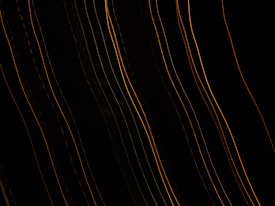 Several curved, parallel lines of yellow and orange light are set against a dark background, creating an abstract, wave-like pattern. The lines vary in brightness and thickness, suggesting motion or dynamic energy.