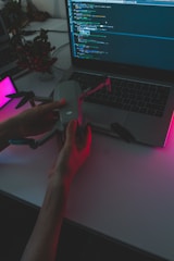 A person holds a drone above a laptop displaying a programming interface. The scene is dimly lit with pink and purple ambient lighting, creating a high-tech and futuristic atmosphere. A potted plant is visible in the background.
