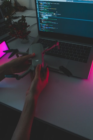 A person holds a drone above a laptop displaying a programming interface. The scene is dimly lit with pink and purple ambient lighting, creating a high-tech and futuristic atmosphere. A potted plant is visible in the background.