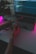 A person holds a drone above a laptop displaying a programming interface. The scene is dimly lit with pink and purple ambient lighting, creating a high-tech and futuristic atmosphere. A potted plant is visible in the background.