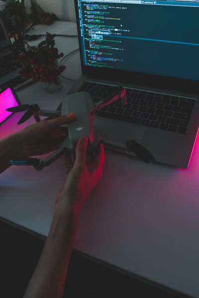 A person holds a drone above a laptop displaying a programming interface. The scene is dimly lit with pink and purple ambient lighting, creating a high-tech and futuristic atmosphere. A potted plant is visible in the background.