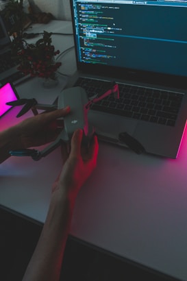 A person holds a drone above a laptop displaying a programming interface. The scene is dimly lit with pink and purple ambient lighting, creating a high-tech and futuristic atmosphere. A potted plant is visible in the background.