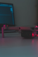 A close-up of a drone resting on a flat surface with a laptop in the background. The scene is dimly lit with soft pink and blue lighting reflecting on the surfaces.