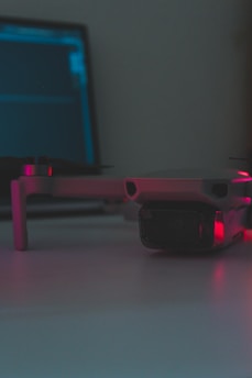 A close-up of a drone resting on a flat surface with a laptop in the background. The scene is dimly lit with soft pink and blue lighting reflecting on the surfaces.