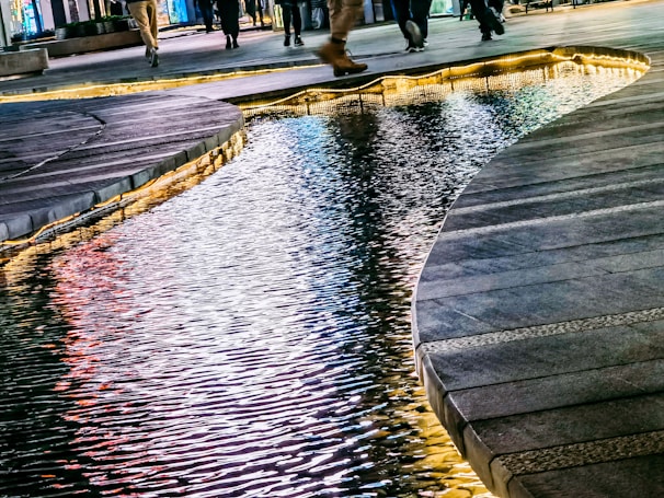 A modern urban scene featuring a reflective body of water seamlessly integrated with the surrounding pavement. The water is lit up with hues of blue and warm colors, creating a vibrant reflection of city lights. Pedestrians are seen walking along the paths, adding a dynamic element to the serene environment.
