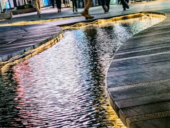 A modern urban scene featuring a reflective body of water seamlessly integrated with the surrounding pavement. The water is lit up with hues of blue and warm colors, creating a vibrant reflection of city lights. Pedestrians are seen walking along the paths, adding a dynamic element to the serene environment.