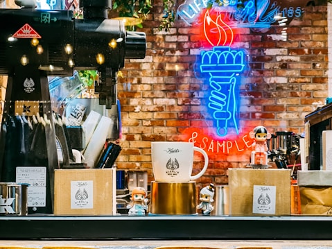A vibrant coffee shop setting with a large coffee grinder positioned on the left. In the foreground, there are Kiehl's branded boxes and a mug, accompanied by small figurines. The background features an illuminated neon sign depicting a hand holding a torch, with the words 'Life Samples' partially visible. The brick wall adds a rustic touch to the scene.