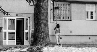 A person is standing in front of a building with the number 49, cleaning or fixing something near a window. The building has a staircase visible through an open door, and a large tree is nearby. The environment looks urban and residential.
