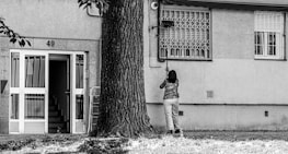 A person is standing in front of a building with the number 49, cleaning or fixing something near a window. The building has a staircase visible through an open door, and a large tree is nearby. The environment looks urban and residential.
