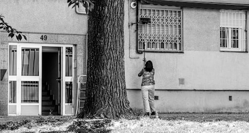 A person is standing in front of a building with the number 49, cleaning or fixing something near a window. The building has a staircase visible through an open door, and a large tree is nearby. The environment looks urban and residential.