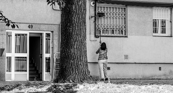 A person is standing in front of a building with the number 49, cleaning or fixing something near a window. The building has a staircase visible through an open door, and a large tree is nearby. The environment looks urban and residential.
