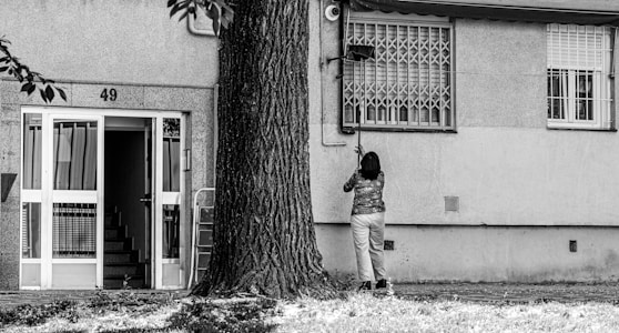 A person is standing in front of a building with the number 49, cleaning or fixing something near a window. The building has a staircase visible through an open door, and a large tree is nearby. The environment looks urban and residential.