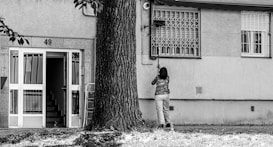 A person is standing in front of a building with the number 49, cleaning or fixing something near a window. The building has a staircase visible through an open door, and a large tree is nearby. The environment looks urban and residential.