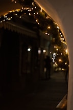 Twinkling string lights decorate a curved archway with pine branches, casting a warm glow in an otherwise dimly lit street scene. Shadows and silhouettes of figures are visible in the background, adding a mysterious ambiance.