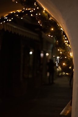 Twinkling string lights decorate a curved archway with pine branches, casting a warm glow in an otherwise dimly lit street scene. Shadows and silhouettes of figures are visible in the background, adding a mysterious ambiance.