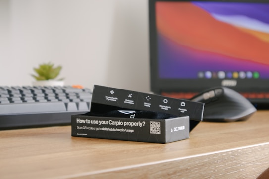 A desk setup featuring a keyboard, computer mouse, a potted plant, and a laptop with a colorful screen. In the foreground, there is a black product box labeled 'How to use your Carpio properly?' The surface of the desk is wooden.