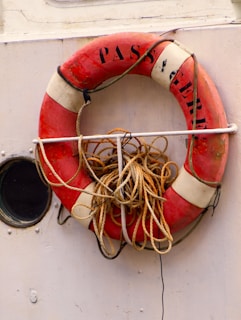A red and white lifebuoy is mounted on the side of a boat with the word 'PASSAGERE' partially visible. It has a bundle of tied ropes attached to it. Next to the lifebuoy is a small round porthole.