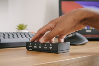A hand is reaching for a small black box placed on a wooden desk. The box has various symbols and text indicating features such as 'minimizes design' and 'portable size'. In the background, there is a computer keyboard, a monitor displaying colorful screens, and a small green plant.