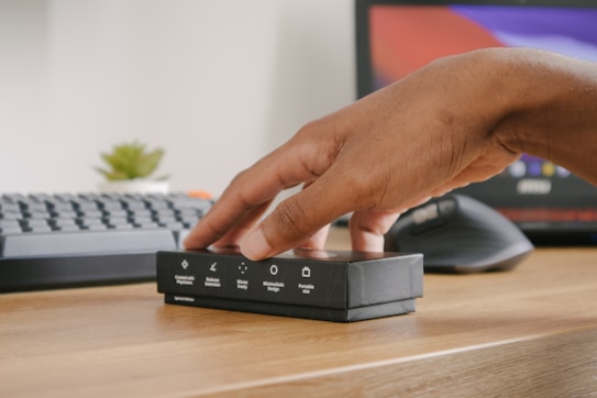 A hand is reaching for a small black box placed on a wooden desk. The box has various symbols and text indicating features such as 'minimizes design' and 'portable size'. In the background, there is a computer keyboard, a monitor displaying colorful screens, and a small green plant.