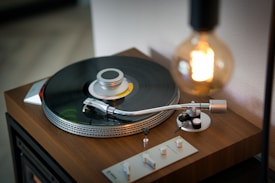 A vintage turntable with a spinning vinyl record is set on a wooden surface, accompanied by a warm glowing light bulb. The turntable arm is positioned over the record, suggesting music is playing. The scene conveys a sense of nostalgia and warmth.
