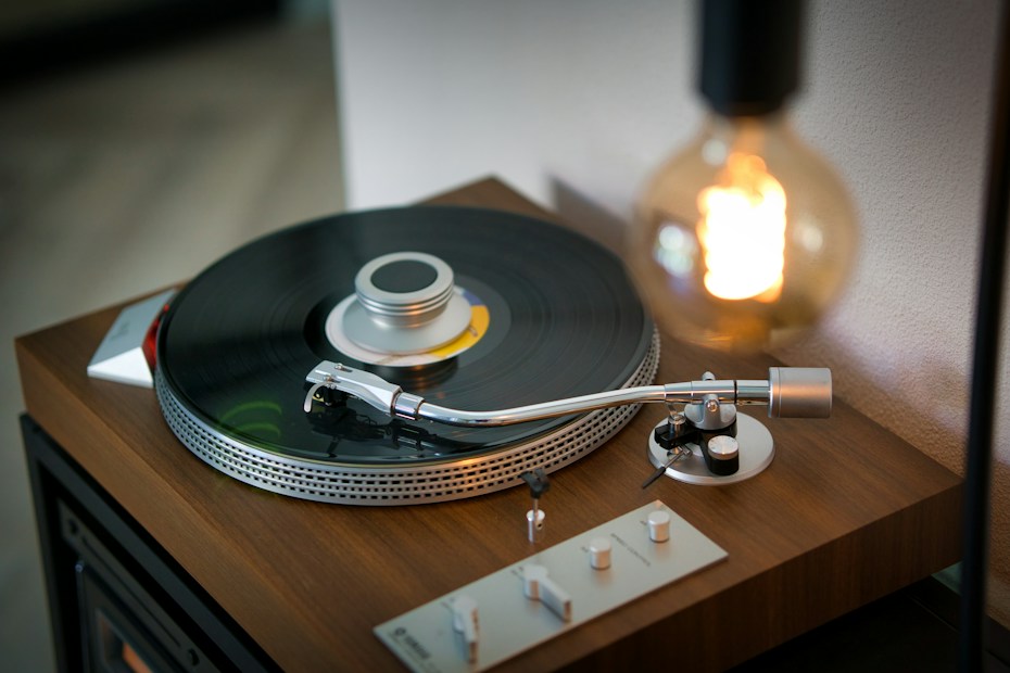 A vintage turntable with a spinning vinyl record is set on a wooden surface, accompanied by a warm glowing light bulb. The turntable arm is positioned over the record, suggesting music is playing. The scene conveys a sense of nostalgia and warmth.