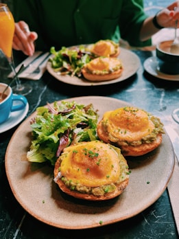Two plates of brunch food featuring eggs benedict-style dishes with creamy sauce and garnished with chives, accompanied by a green salad. Drinking glasses and cups are visible on a dark marble table surface, with two people partaking in the meal.