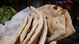 Several pieces of baked flatbread, possibly naan, are stacked in a basket. The bread has a slightly charred appearance, with a soft texture. In the background, there are some green leaves, possibly herbs or vegetables.