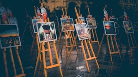 A series of easels display framed photographs, arranged neatly in an outdoor setting. The lighting is dim, creating a moody atmosphere, possibly suggesting an evening exhibition. The ground appears wet, reflecting the easels and photographs, adding to the ambiance. Small decorative figures are affixed at the top of each easel.