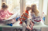 Three children are sitting on a windowsill. One child is reading a large book while the other two look outside the window. They are dressed in cozy, colorful sweaters and pajamas, and the atmosphere outside suggests a cold, wintry day. A couple of toys and another book are placed nearby.
