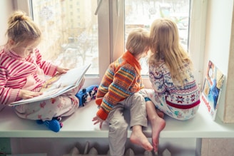 Three children are sitting on a windowsill. One child is reading a large book while the other two look outside the window. They are dressed in cozy, colorful sweaters and pajamas, and the atmosphere outside suggests a cold, wintry day. A couple of toys and another book are placed nearby.