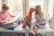 Three children are sitting on a windowsill. One child is reading a large book while the other two look outside the window. They are dressed in cozy, colorful sweaters and pajamas, and the atmosphere outside suggests a cold, wintry day. A couple of toys and another book are placed nearby.