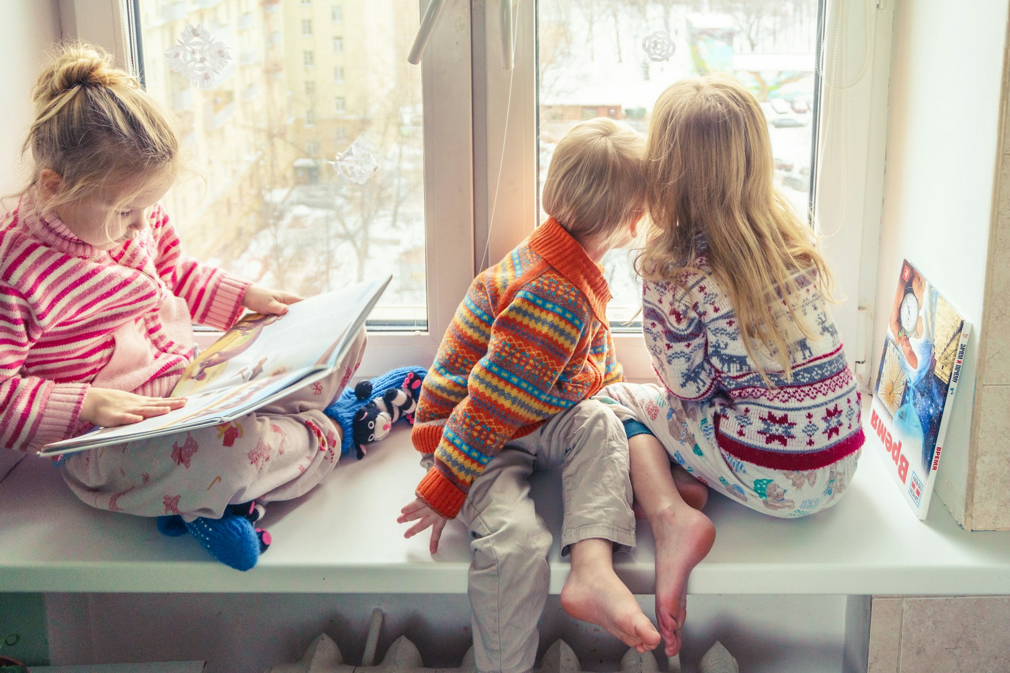 a group of children with blonde hair in knitted sweatshirts with northern patterns sit near the window, and it's winter outside