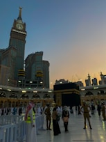 A view of the Kaaba at the Grand Mosque, surrounded by worshippers and pilgrims. The Abraj Al Bait Clock Tower is visible in the background, towering over the mosque complex. The sky is transitioning from day to night, with a blend of sunset colors.