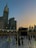 A view of the Kaaba at the Grand Mosque, surrounded by worshippers and pilgrims. The Abraj Al Bait Clock Tower is visible in the background, towering over the mosque complex. The sky is transitioning from day to night, with a blend of sunset colors.